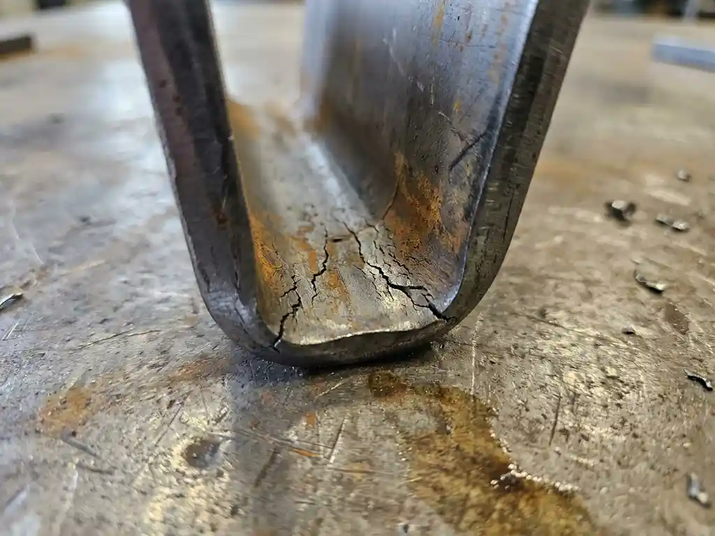 Gebogen staalplaat met scheurvorming en spanningsfracturen langs de binnenradius op een industriële stalen werkbank.